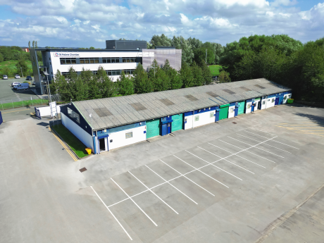 A long, single-storey industrial building with blue doors is adjacent to an empty car park; an office block and trees are in the background under a partly cloudy sky.