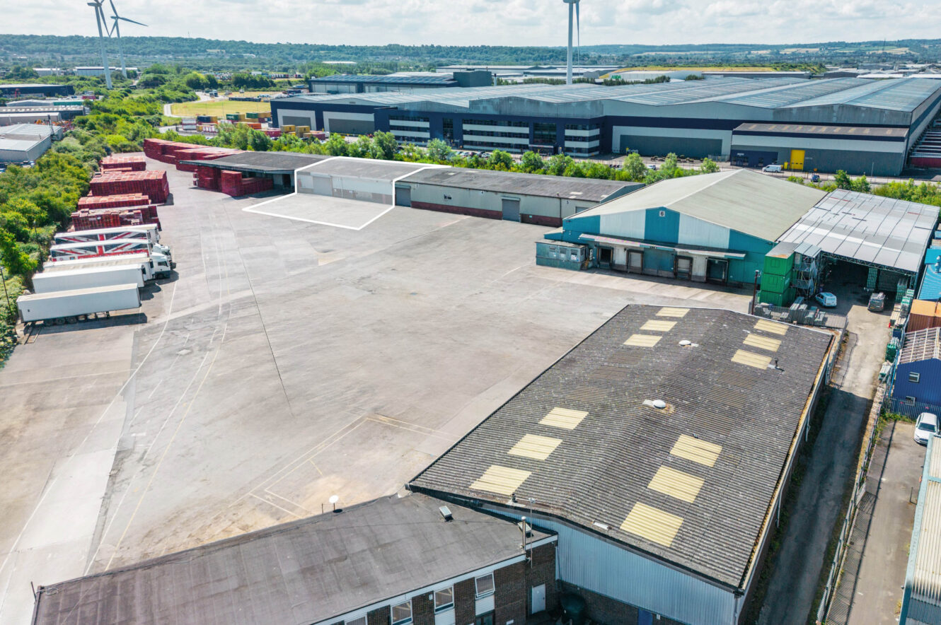 Aerial view of an industrial facility with warehouses, shipping containers, lorries, and large paved areas on a sunny day.