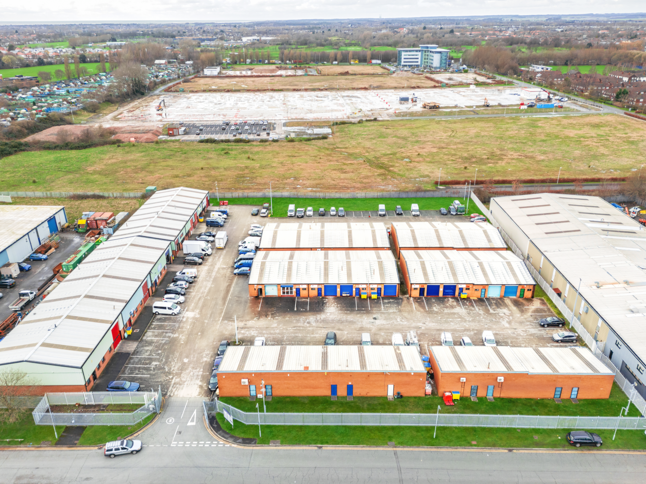 Aerial view of industrial warehouses with car parks, surrounded by open fields and construction area in the background.