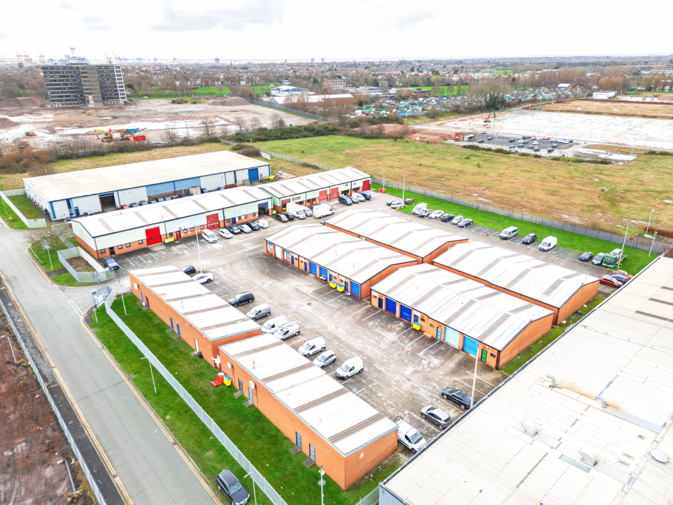 Aerial view of an industrial estate with several warehouse units, parked cars, and surrounding open land under a cloudy sky.