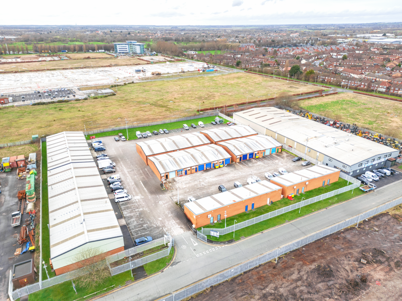 Aerial view of an industrial estate with multiple warehouse buildings, parked vehicles, and adjacent open fields and residential areas in the background.