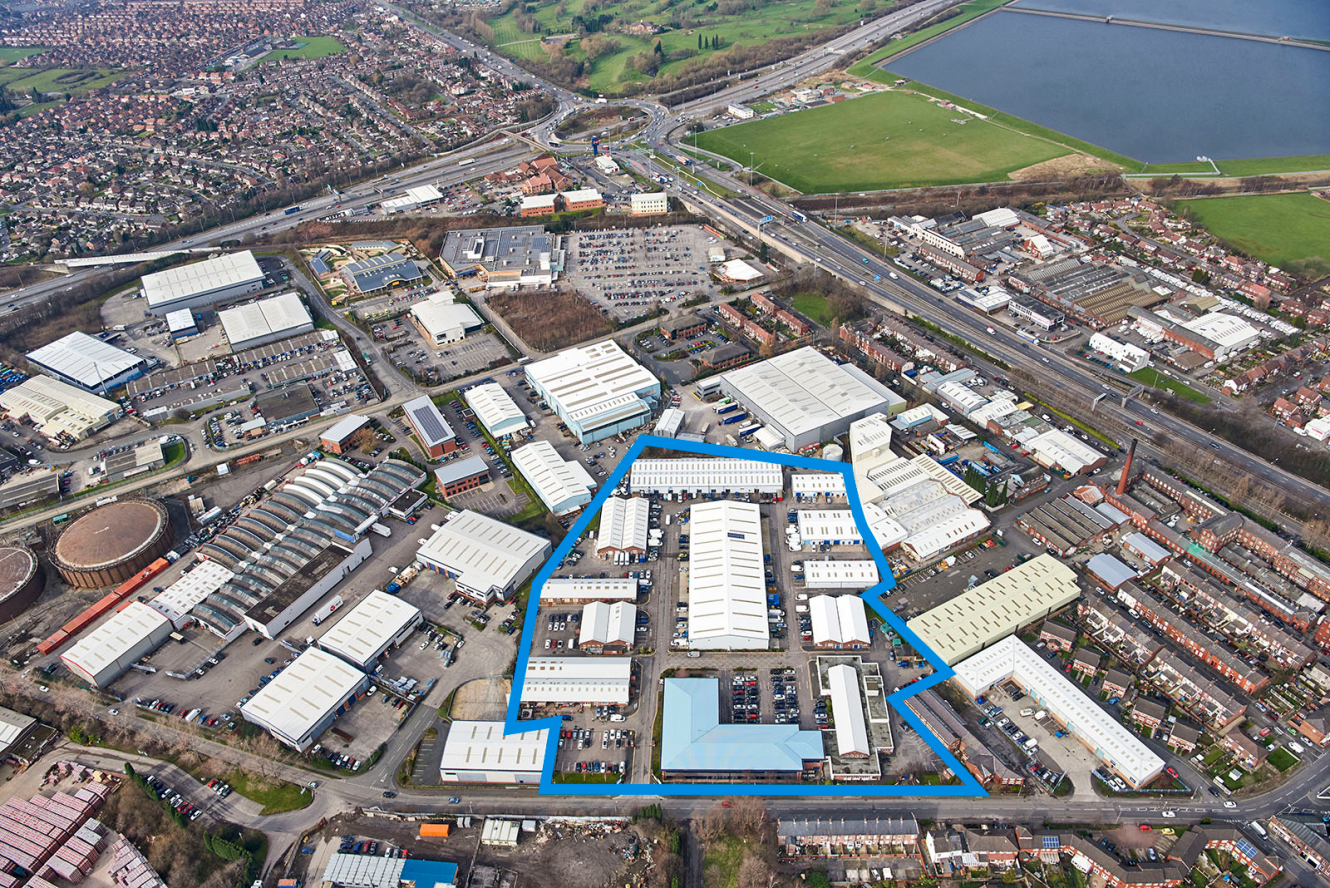 An aerial view of an industrial estate with large white-roofed buildings, outlined in blue, surrounded by roads, residential areas, and a reservoir.