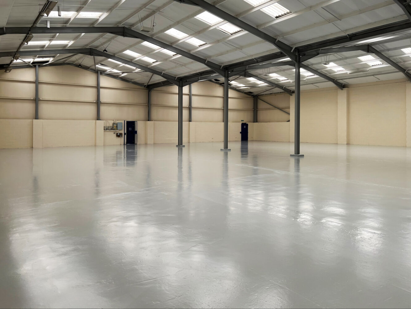 Empty, spacious industrial warehouse with a shiny, light grey floor, high ceiling, exposed beams, beige walls, and blue doors. Fluorescent lights are mounted on the ceiling.