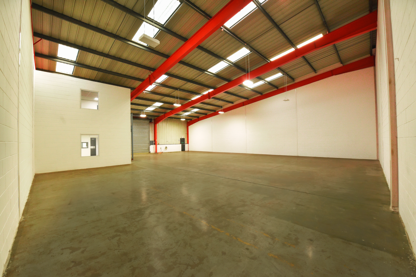 Interior of an empty warehouse with concrete floors, high ceilings, red roof beams, white walls, and a small office area on the left.