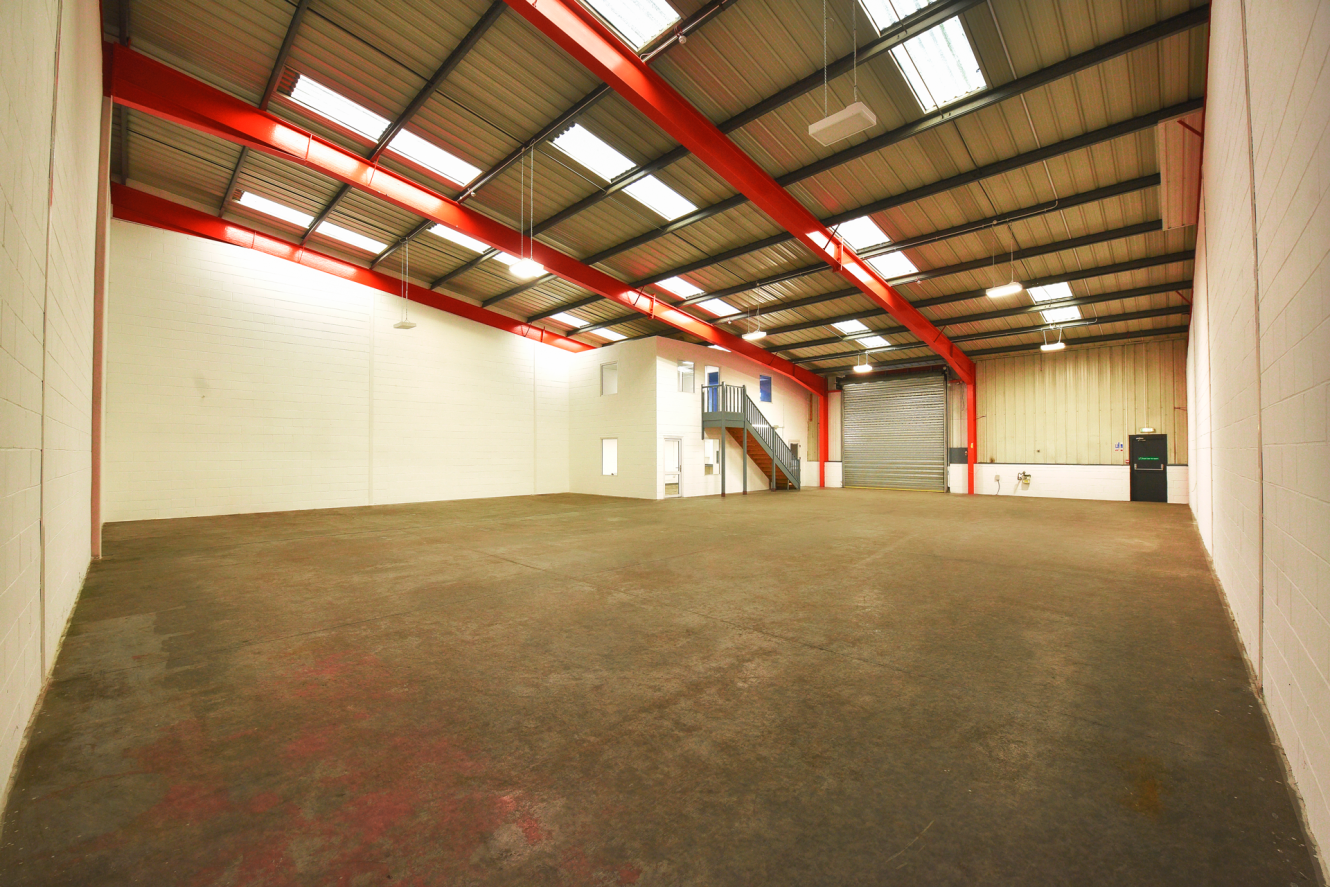 Interior of a large, empty warehouse with red steel beams, high ceilings, concrete floor, white walls, and a staircase leading to a small office area.
