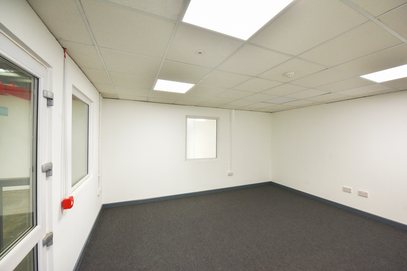 An empty office room with white walls, grey carpet, ceiling lights, and three windows; electrical sockets are visible on one wall.