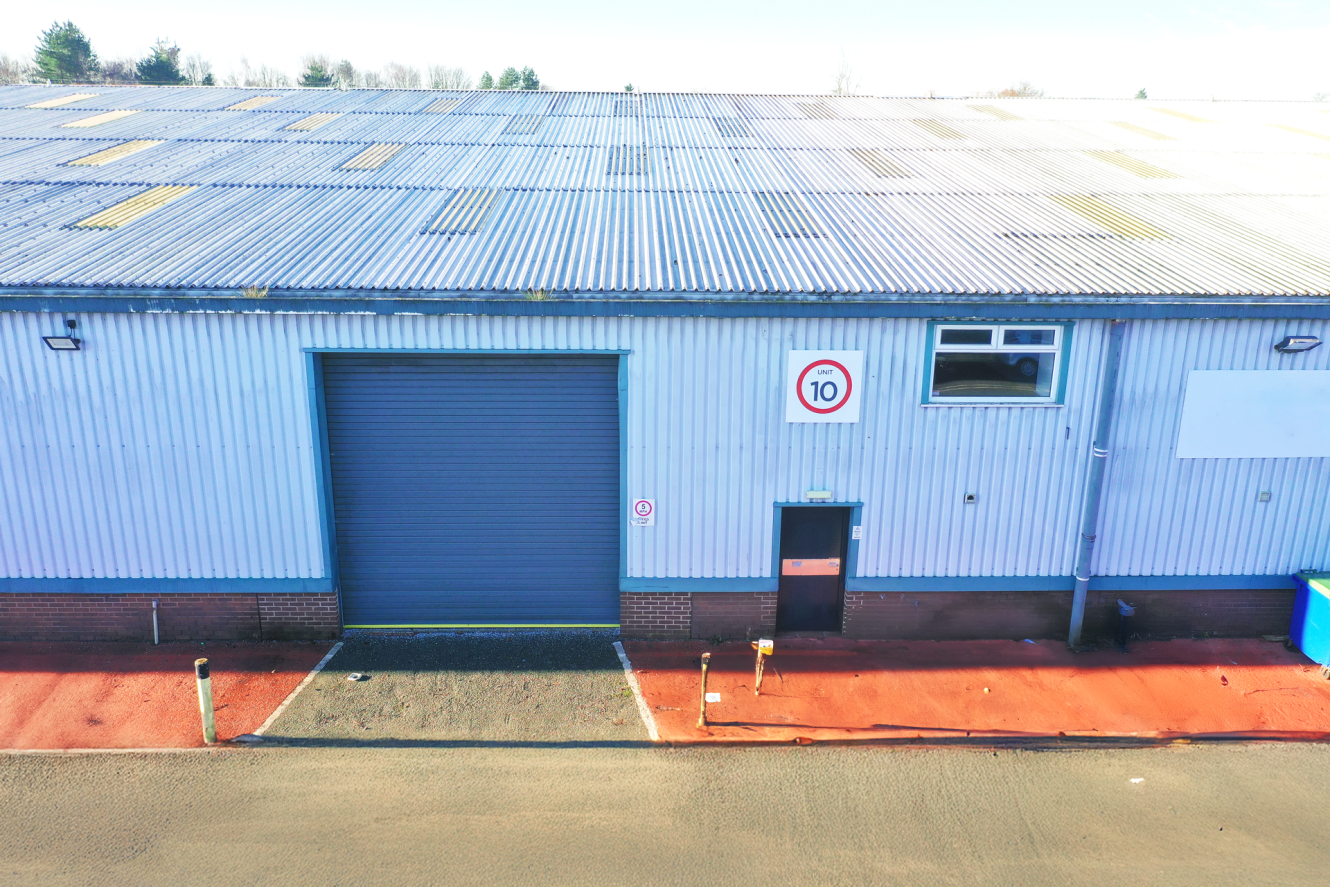 Industrial warehouse exterior with a large closed roller shutter, a small entrance door, a 10 mph speed limit sign, and a blue bin on red-painted pavement.