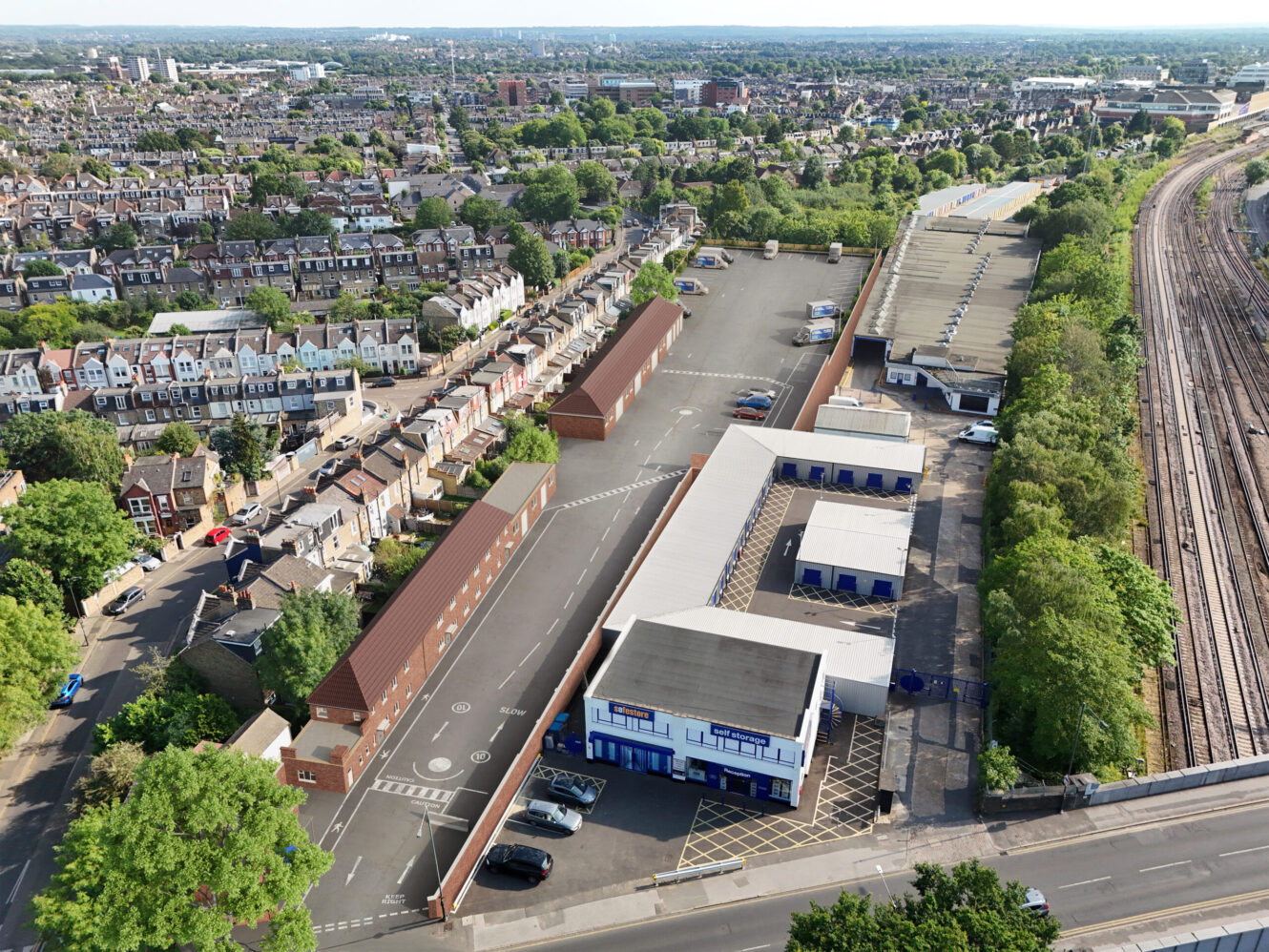 Aerial view of an industrial estate with warehouses, car parks, and nearby residential houses, adjacent to railway tracks and a green area.
