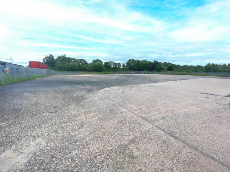 Large empty concrete car park with some worn and darkened areas, bordered by a metal fence on the left and trees in the background under a partly cloudy sky.