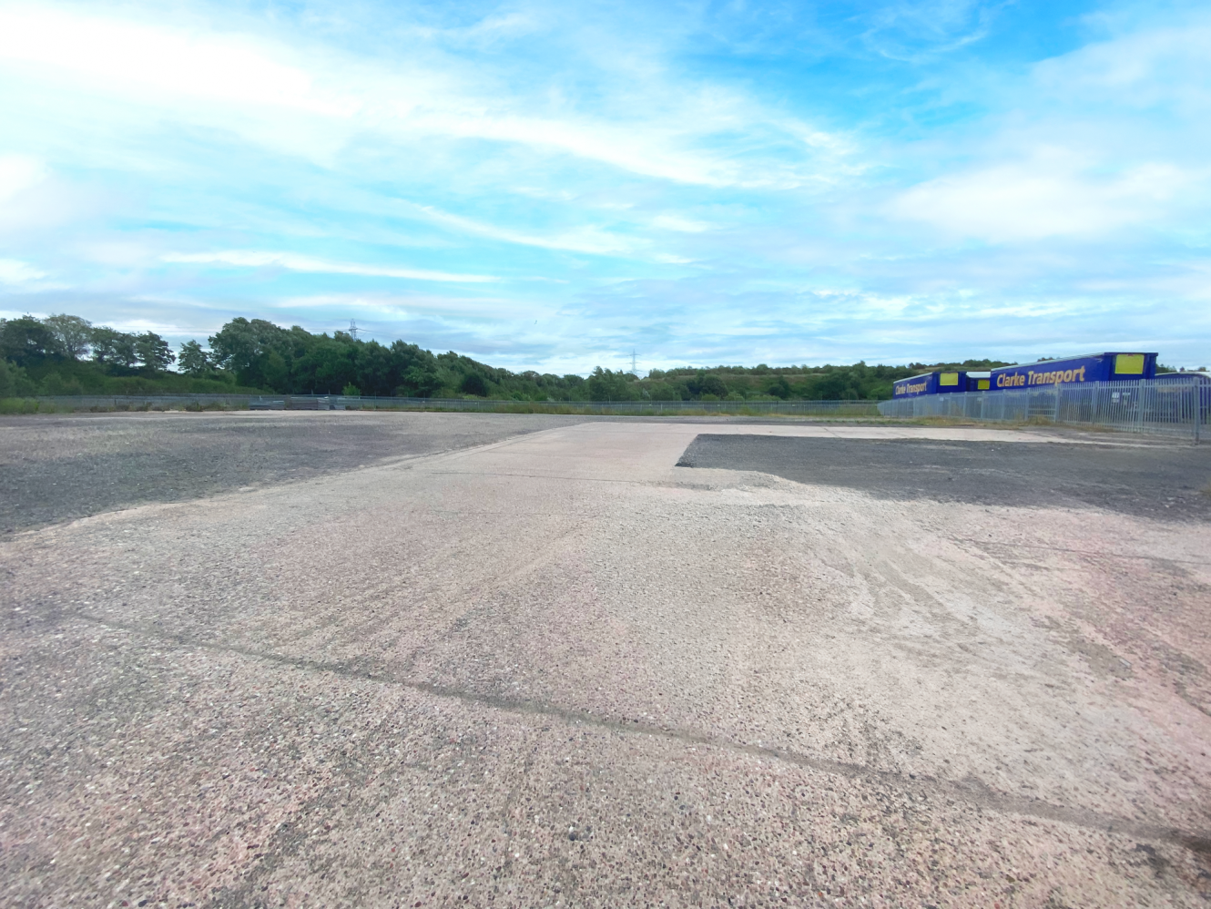 An empty, paved car park with some gravel areas, bordered by trees. A blue building with C Saffron Transport signage is visible on the right.
