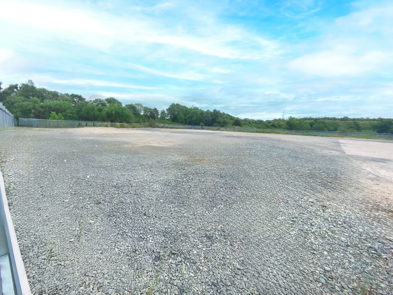 A large, empty gravel car park bordered by a metal fence and surrounded by trees under a partly cloudy sky.