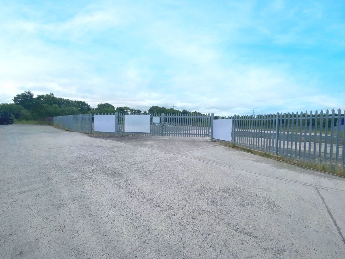 A wide paved area leads to a closed metal security gate with blank white signs, surrounded by fencing and greenery under a partly cloudy sky.