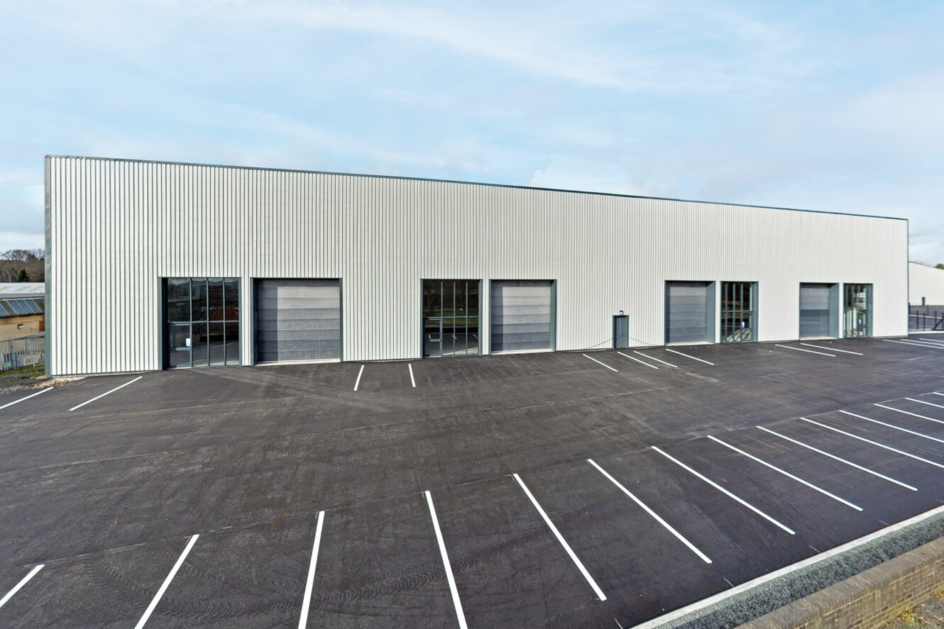 A large industrial warehouse with four loading bays and several glass doors, set beside an empty car park with marked spaces.