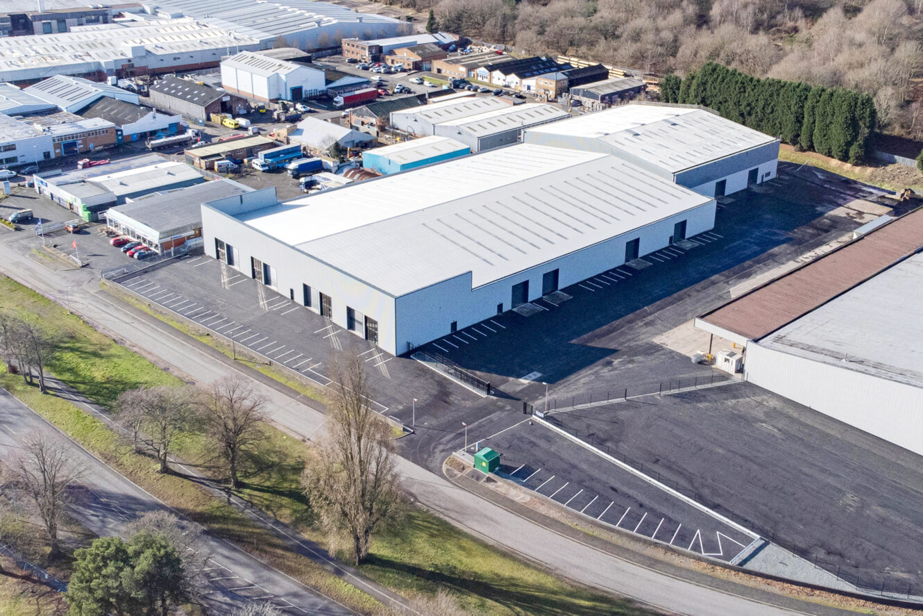 Aerial view of a large industrial warehouse with white roof, surrounded by car parks and other commercial buildings, adjacent to a tree-lined road.