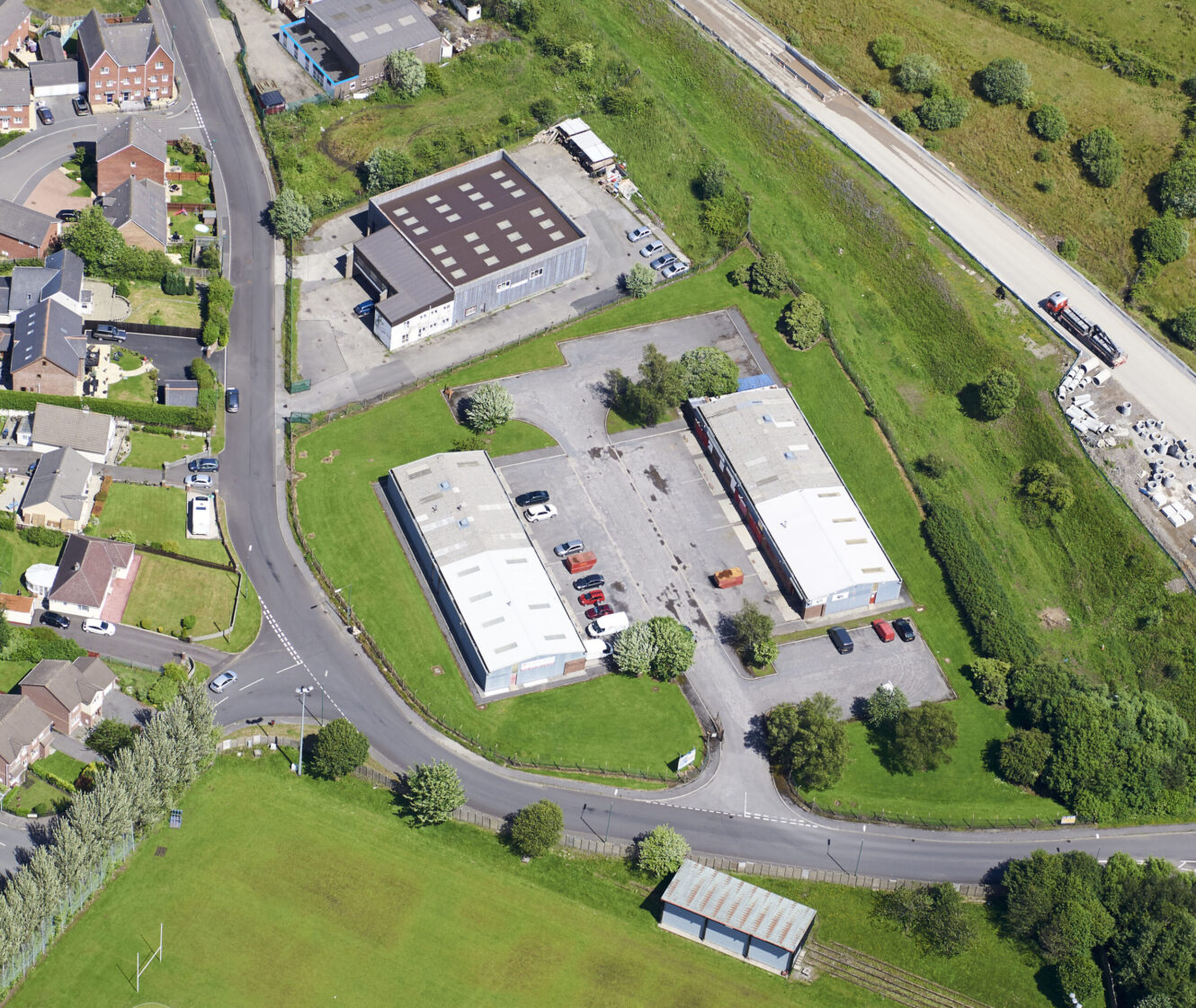 Aerial view of two industrial buildings with car parks, surrounded by grass, trees, nearby houses, and a road with vehicles.