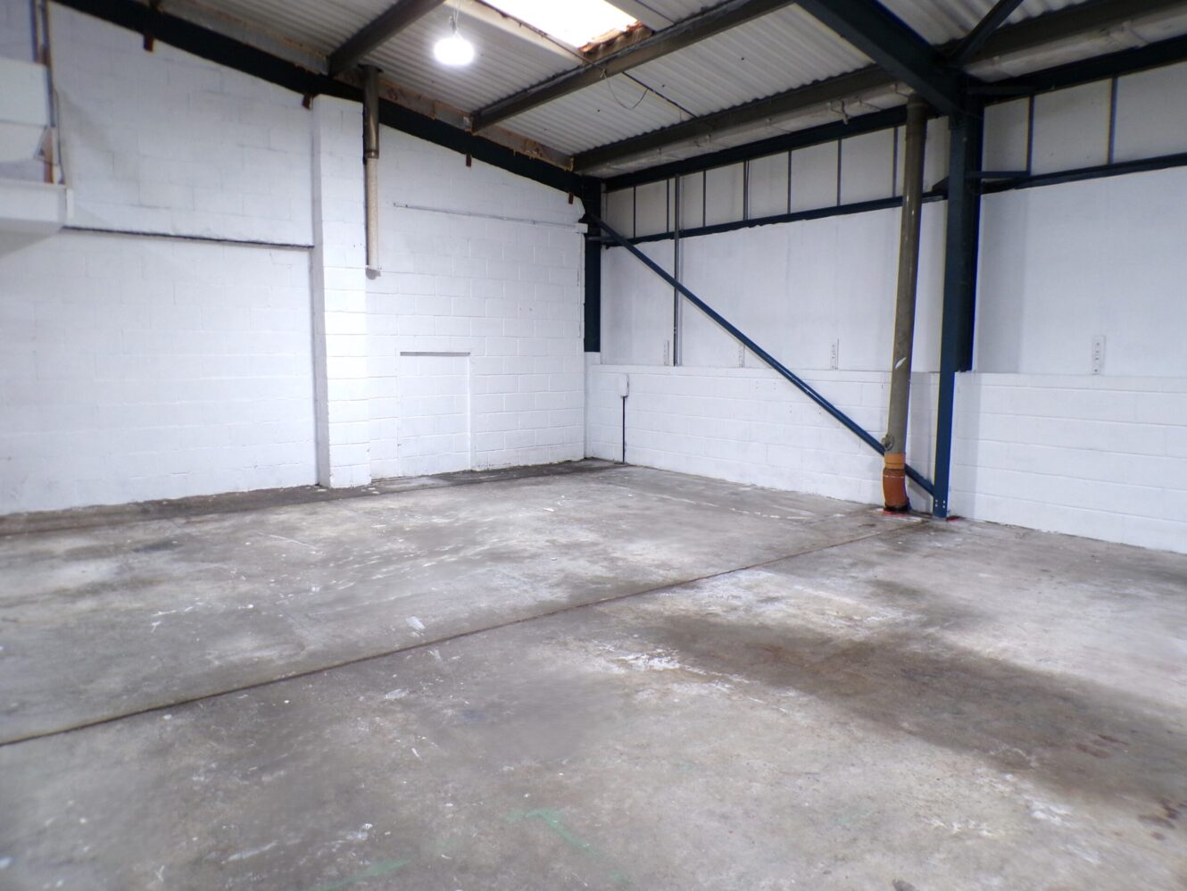 Empty industrial warehouse with concrete floor, white painted brick walls, exposed beams, and a high ceiling with a single overhead light.