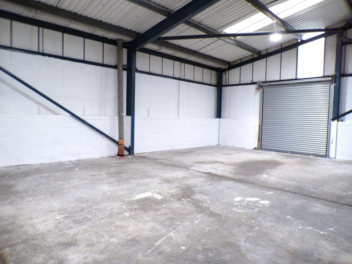 Empty industrial warehouse interior with concrete floor, white painted walls, exposed steel beams, and a partially open metal roller shutter.