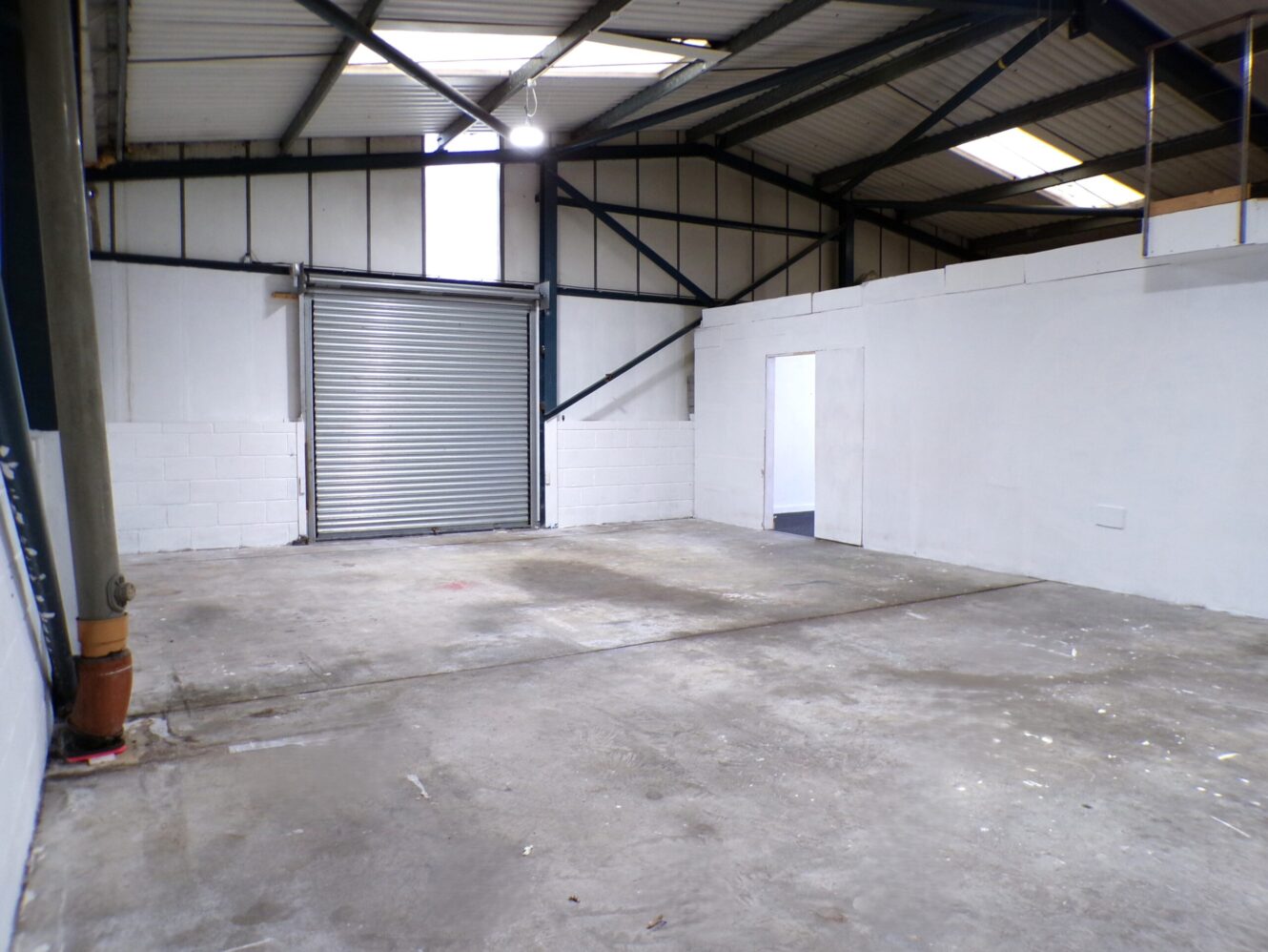 Empty industrial warehouse interior with concrete floor, white walls, metal ceiling, skylights, a roller shutter door, and an open doorway leading to another room.