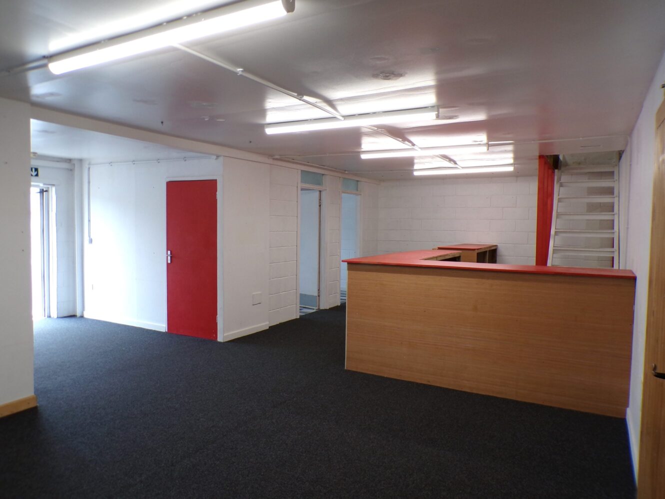 An empty office space with a wooden reception desk, red accents on doors and counters, fluorescent lighting, and a staircase in the corner.