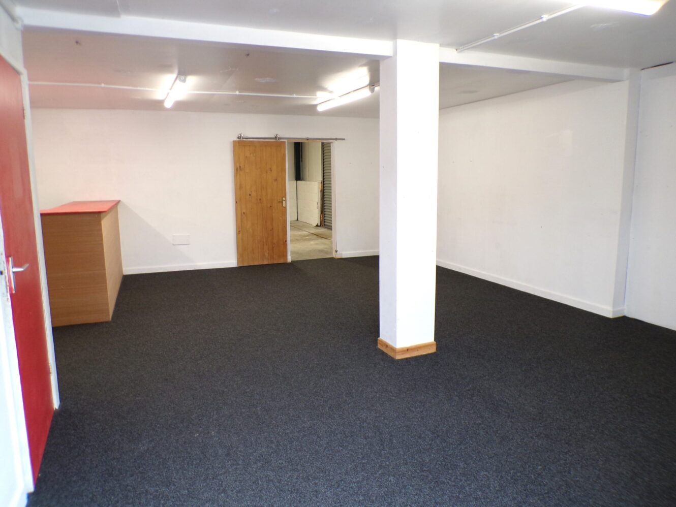 Unfurnished room with white walls, dark carpet, a wooden counter, and a wooden door leading to another area. Fluorescent ceiling lights are on.