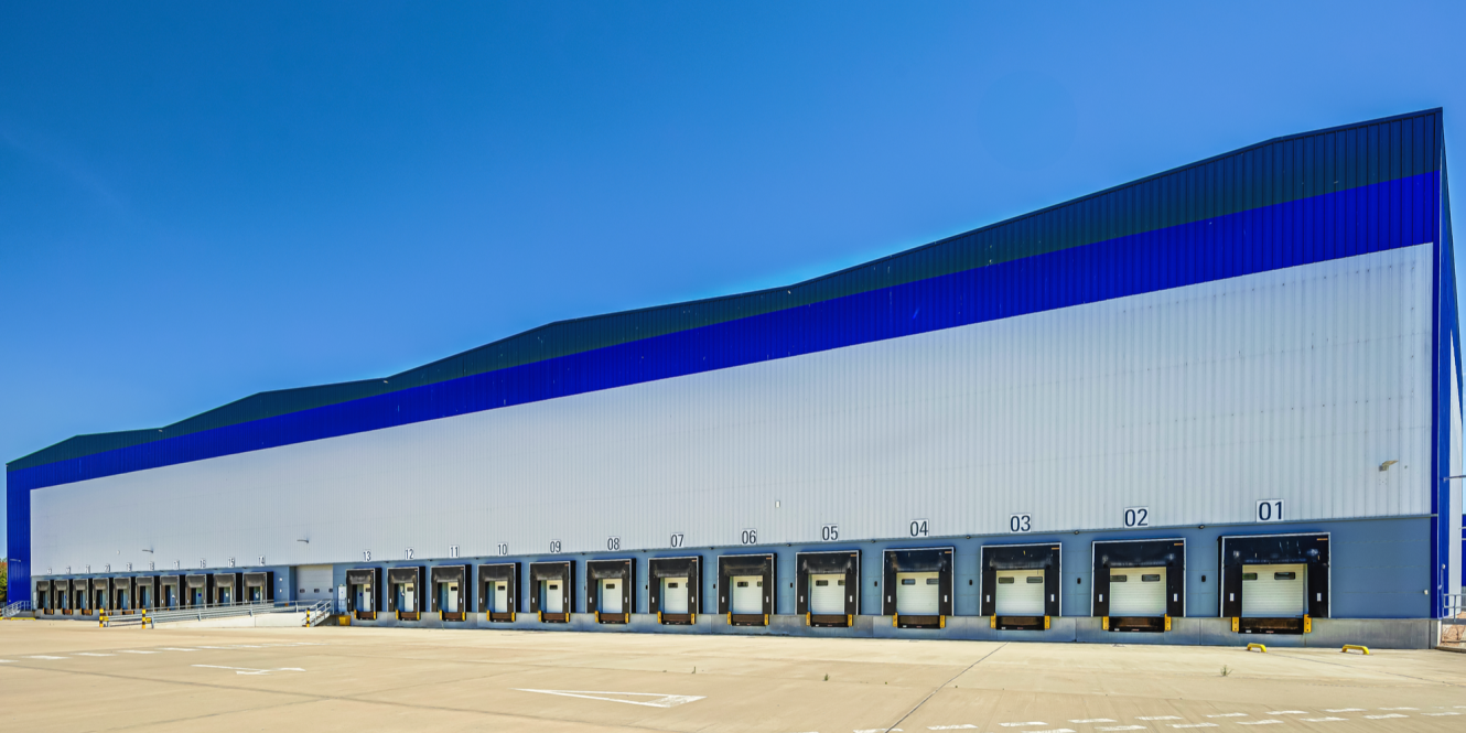 A large industrial warehouse with numbered loading bay doors and a blue and white exterior under a clear sky.