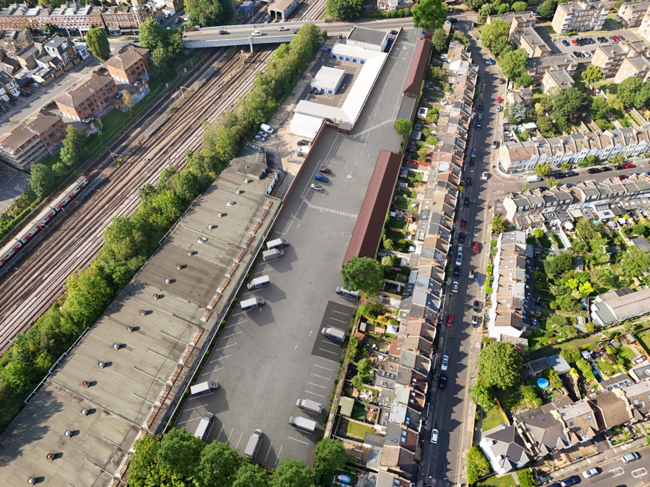 Aerial view of a railway, adjacent car park with several vehicles, and nearby residential houses lined along a street.