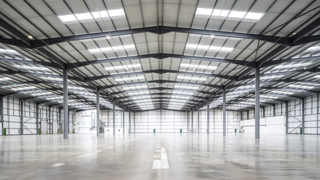 Spacious, empty warehouse interior with high ceilings, metal beams, and polished concrete floors under a roof of large rooflights.