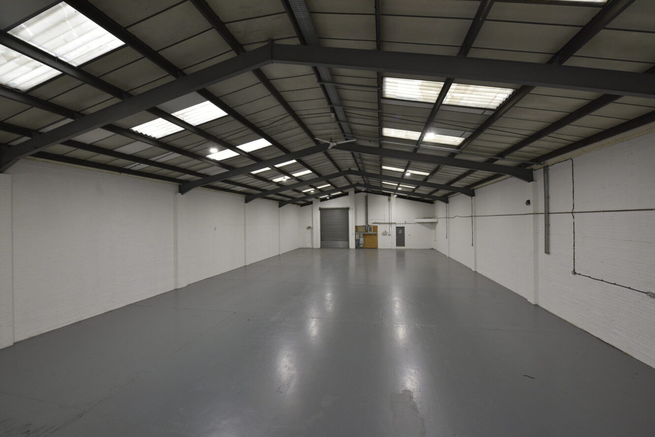 Interior of an empty industrial warehouse with white painted brick walls, a grey concrete floor, and a metal beam ceiling with skylights. A large roller shutter is visible at the far end.