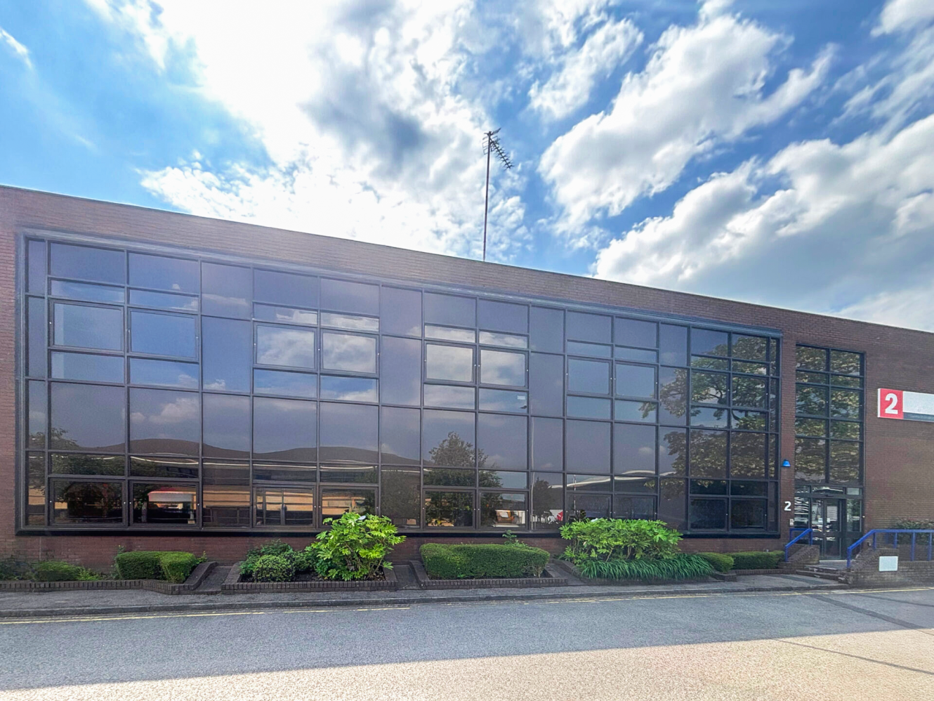 A modern two-storey brick office building with large glass windows, green shrubs in front, and a sign with the number 2 near the entrance on a partly cloudy day.