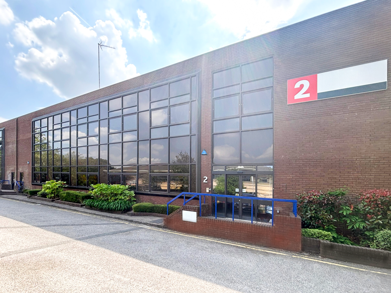 A modern brick industrial building with large glass windows, a blue railing, green plants, and a red sign with the number 2 near the entrance.