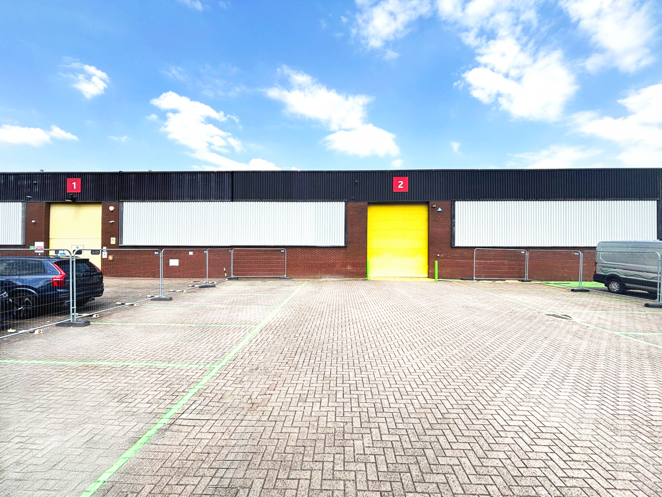 A brick warehouse with two large yellow doors labelled 1 and 2, fenced car park, and some parked vehicles under a partly cloudy sky.