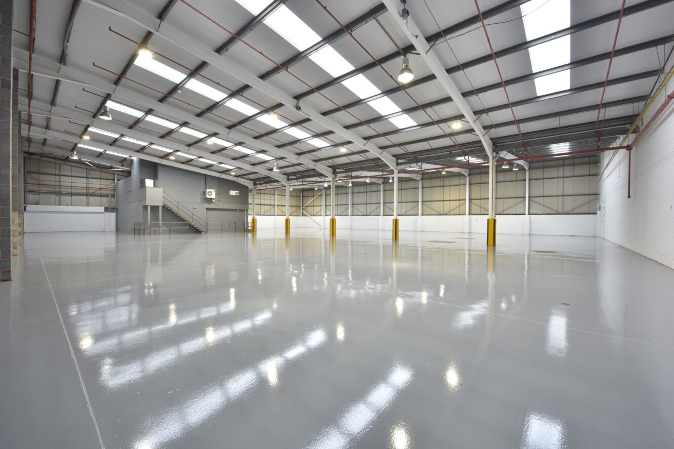 Large, empty warehouse with polished concrete floors, white walls, high ceilings, and exposed beams. A staircase leads to a mezzanine level at the far end.