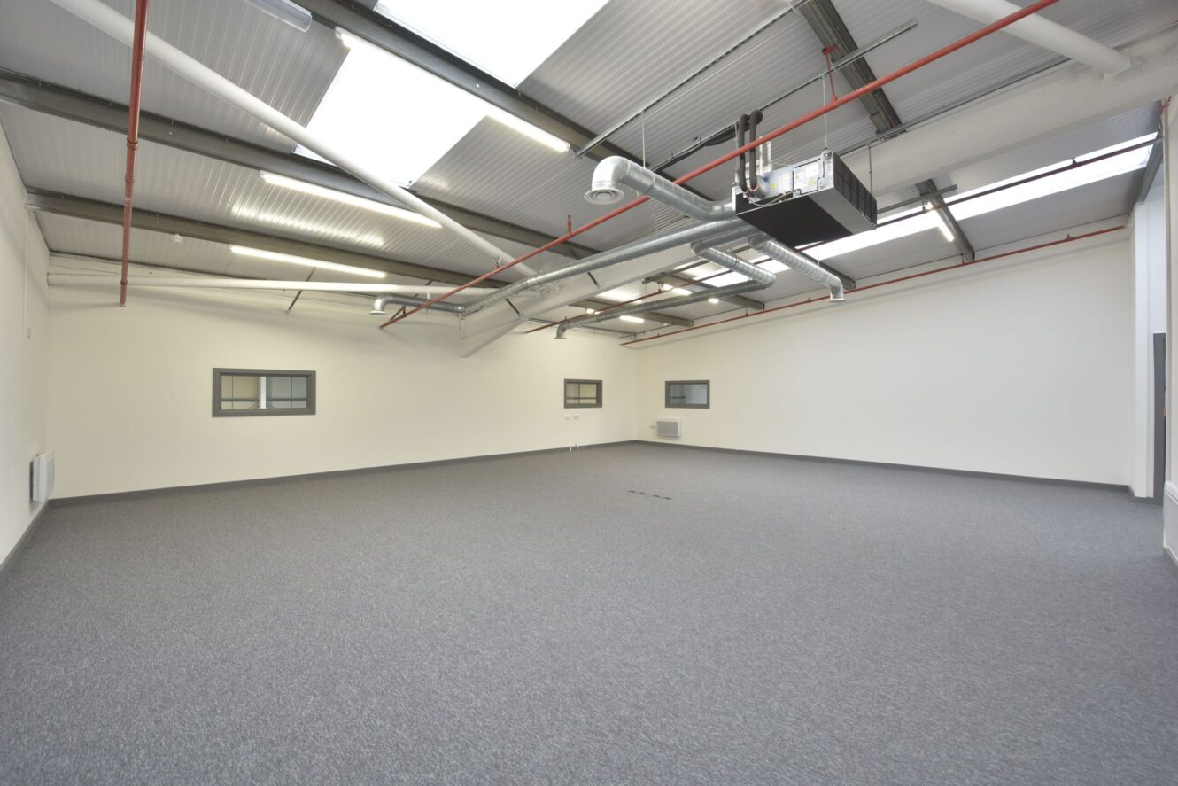 Empty office space with grey carpet, white walls, several small windows, and exposed ceiling pipes and ductwork under angled skylights.