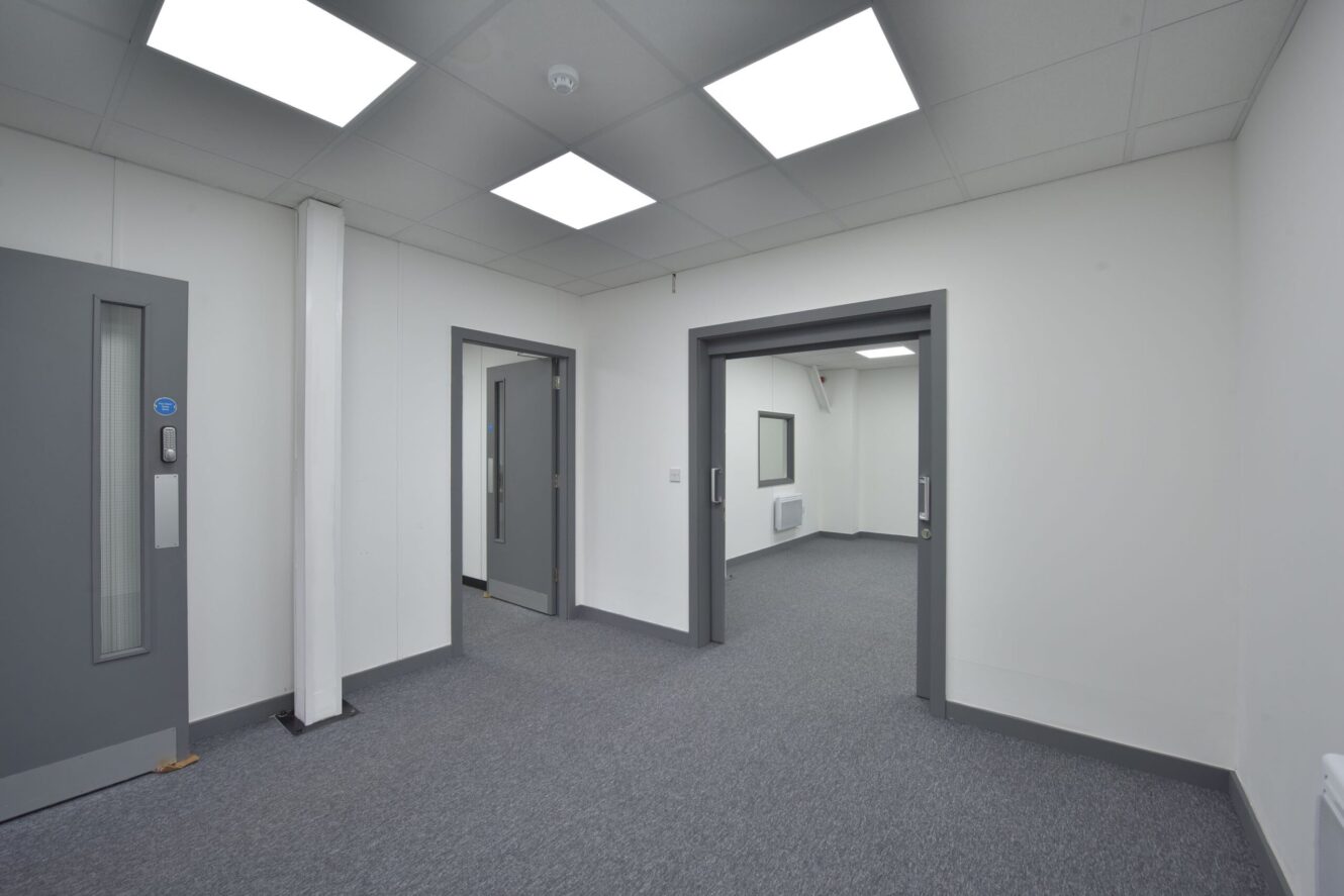 A modern, empty office space with grey carpet, white walls, and several open doorways leading to adjoining rooms. Fluorescent ceiling lights illuminate the area.