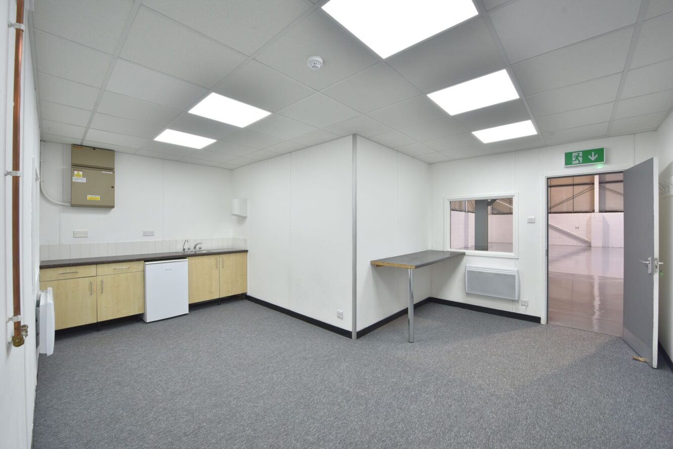 A small, empty office room with grey carpet, white walls, a kitchenette, a counter, fluorescent ceiling lights, and an open door leading to a warehouse area.