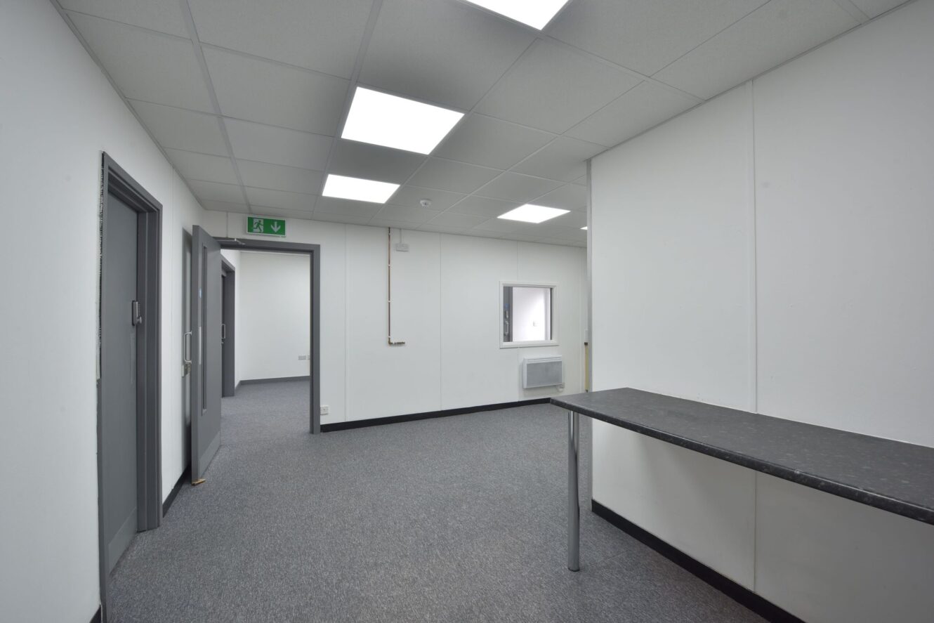 A clean, empty office corridor with grey carpet, white walls, several closed doors, fluorescent ceiling lights, and a long black worktop on the right.