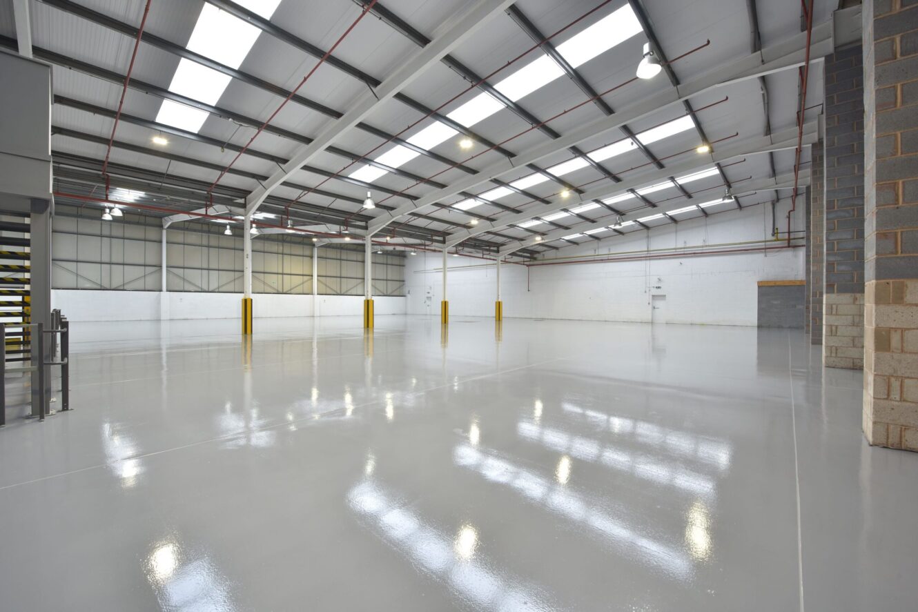 A large, empty warehouse interior with a polished concrete floor, high ceiling, exposed beams, and several yellow bollards.
