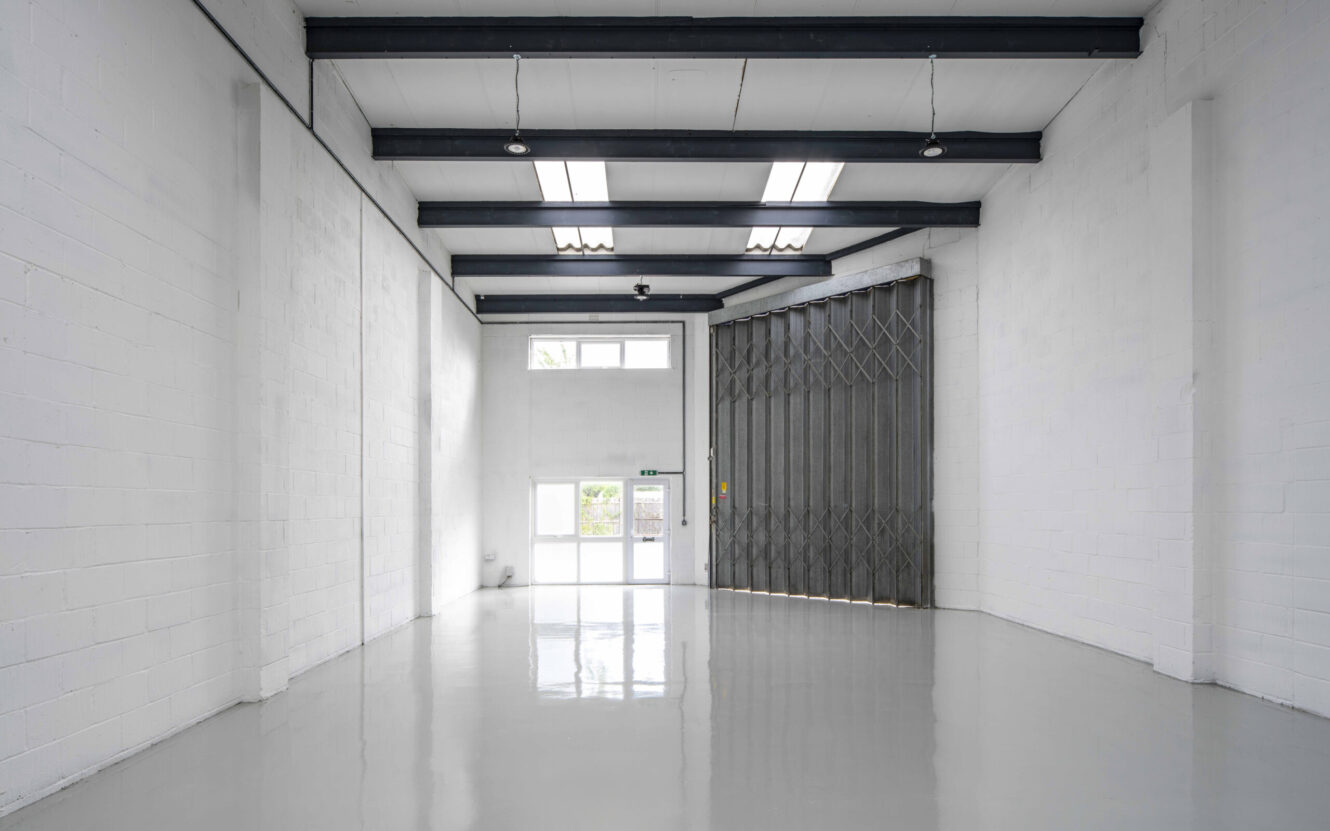 Empty industrial warehouse unit with white walls, high ceiling, polished grey floor, metal beams, large folding metal door, and a single glass exit door at the back.