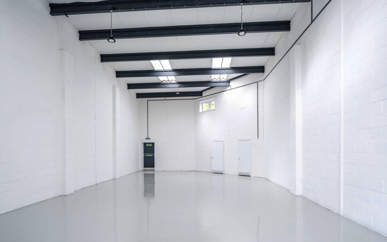 Empty industrial warehouse with white painted brick walls, polished grey floor, exposed ceiling beams, fluorescent lighting, and a green exit door at the back.