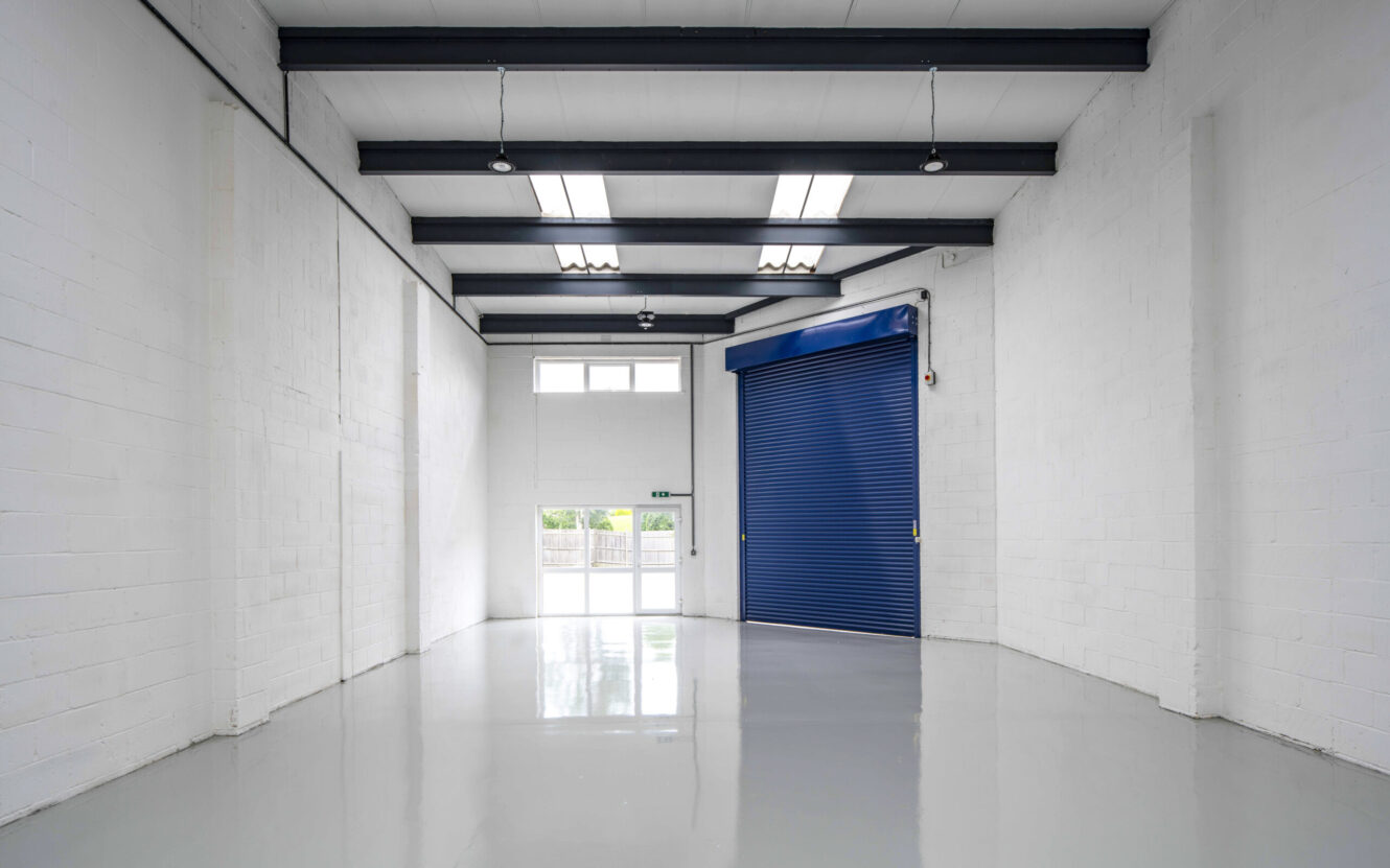 Empty industrial warehouse with white walls, a polished grey floor, ceiling lights, and a large blue roller shutter door. Natural light enters through windows and a glass door.