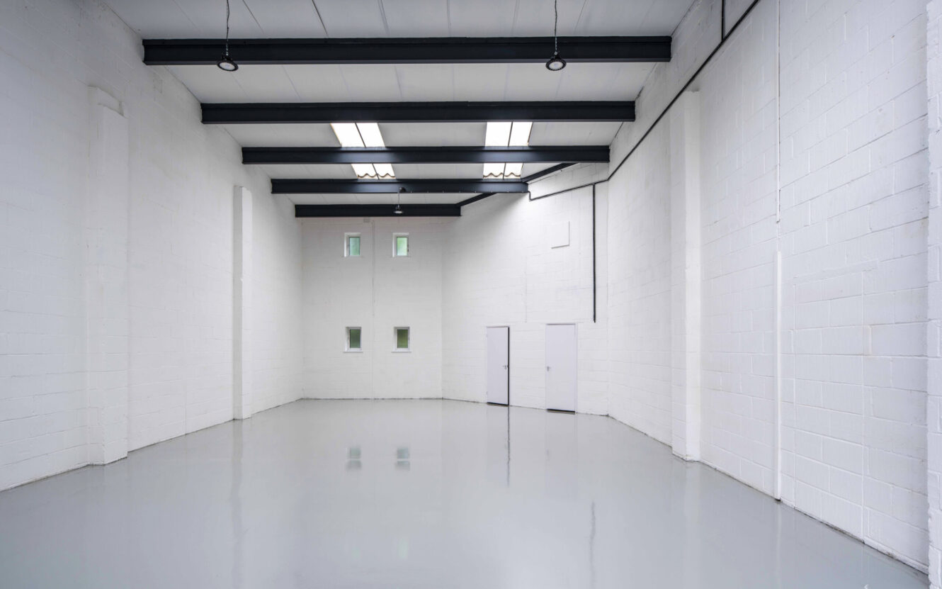Empty industrial warehouse with white brick walls, glossy grey floor, ceiling beams, windows, and two closed doors at the far end.