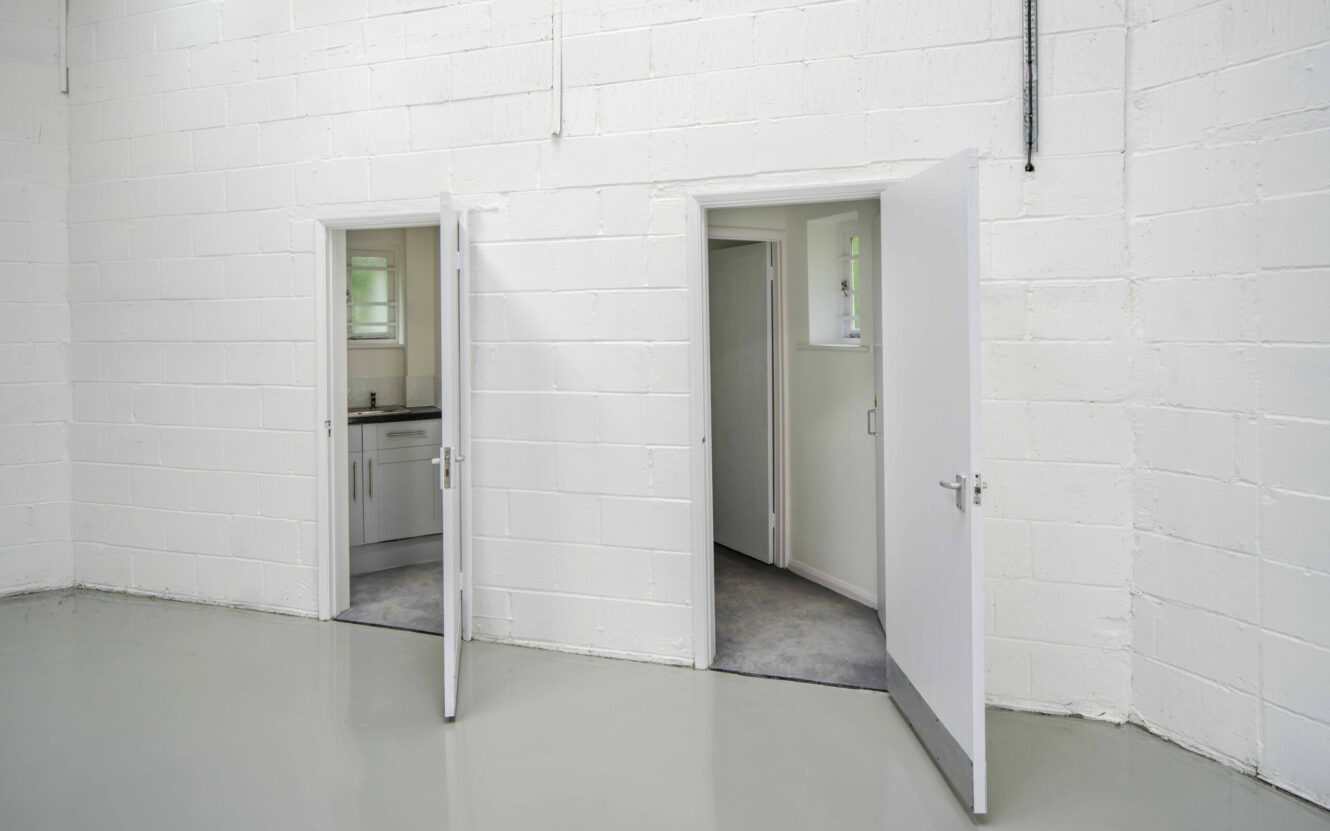 Two open white doors in a white-painted brick wall reveal small rooms, one containing a kitchenette with cupboards and a sink, in an empty, clean, industrial space.