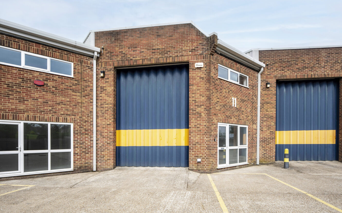 A modern industrial unit with a large blue and yellow roller shutter door, brick exterior, glass windows, and a paved car park in front.