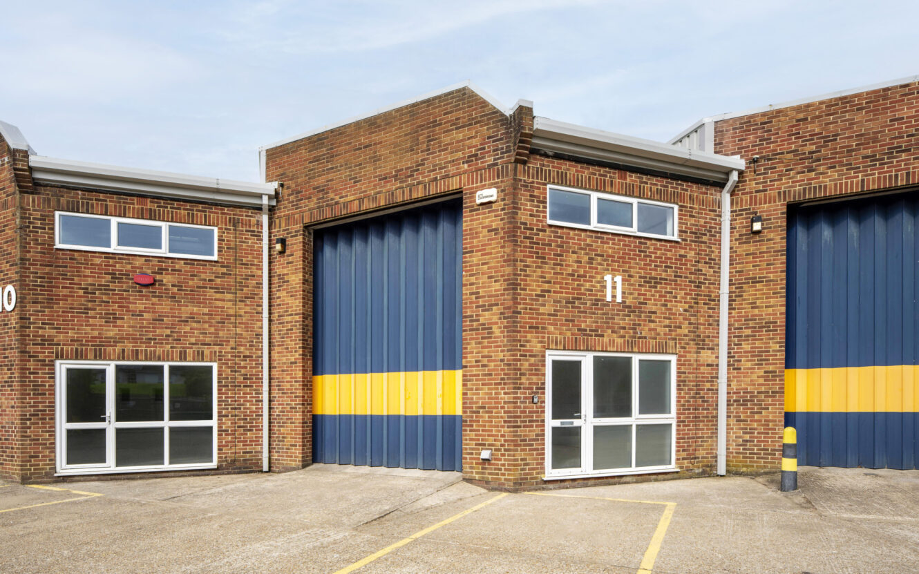 A brick industrial unit with a large blue and yellow roller shutter door, labelled 11, and two glass entrance doors, in an empty car park.