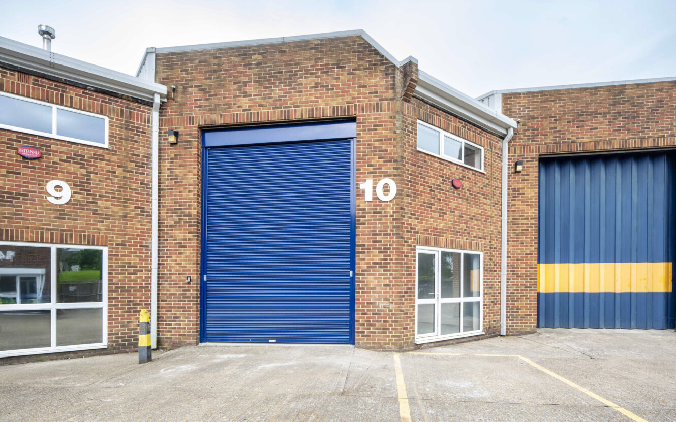 Brick industrial building with a blue roller shutter door numbered 10, adjacent to units 9 and 11, and parking spaces in front.
