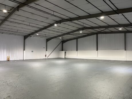 An empty warehouse with a grey concrete floor, white panelled walls, and exposed steel beams under a high ceiling with bright overhead lights.