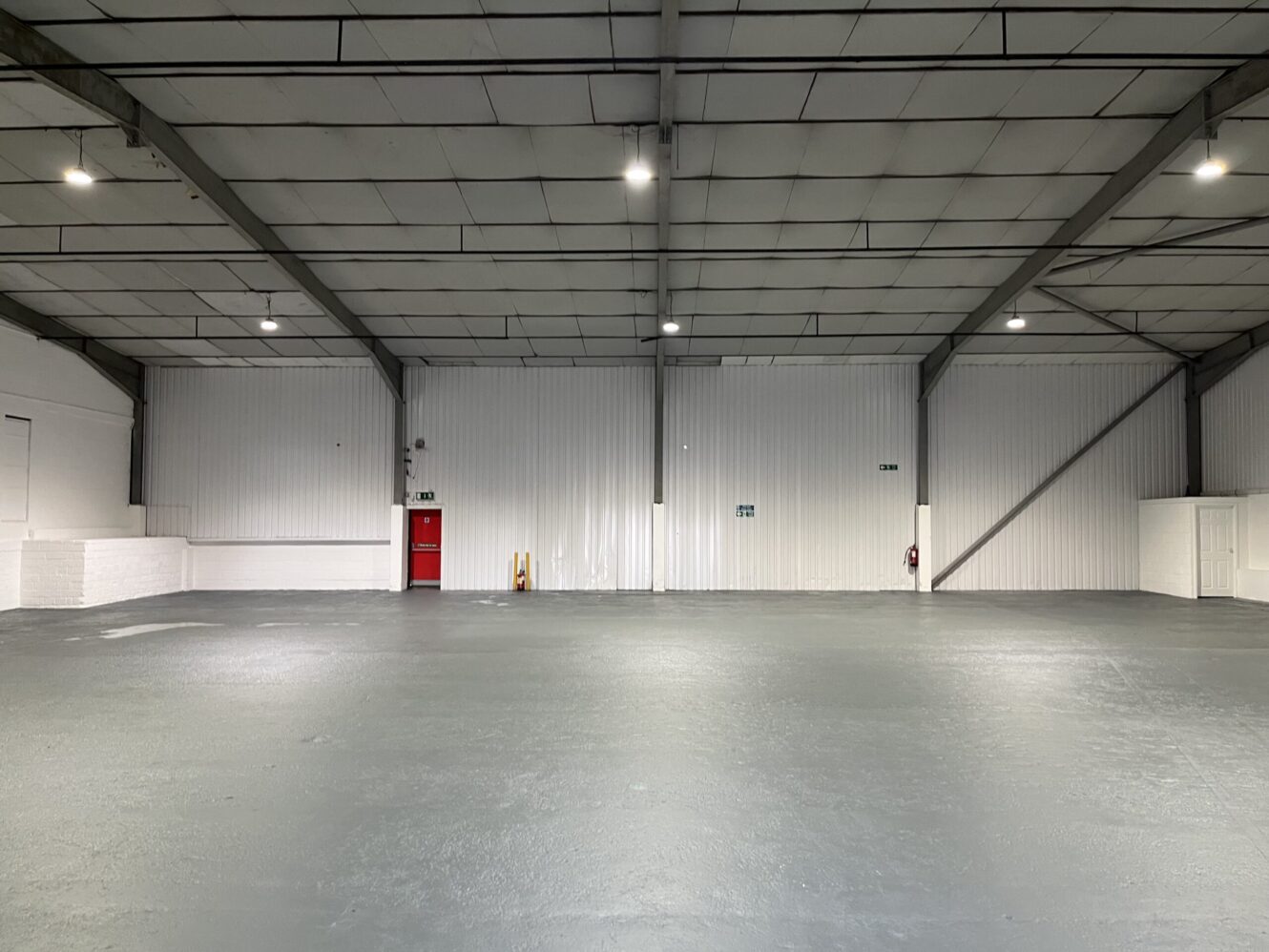 Empty industrial warehouse with white walls, grey floor, exposed ceiling beams, fluorescent lighting, red emergency exit door, and fire safety equipment along the back wall.