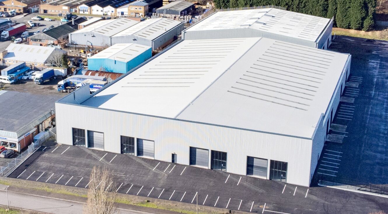 An aerial view of a large, modern warehouse building with multiple loading bays and a nearly empty car park in an industrial area.