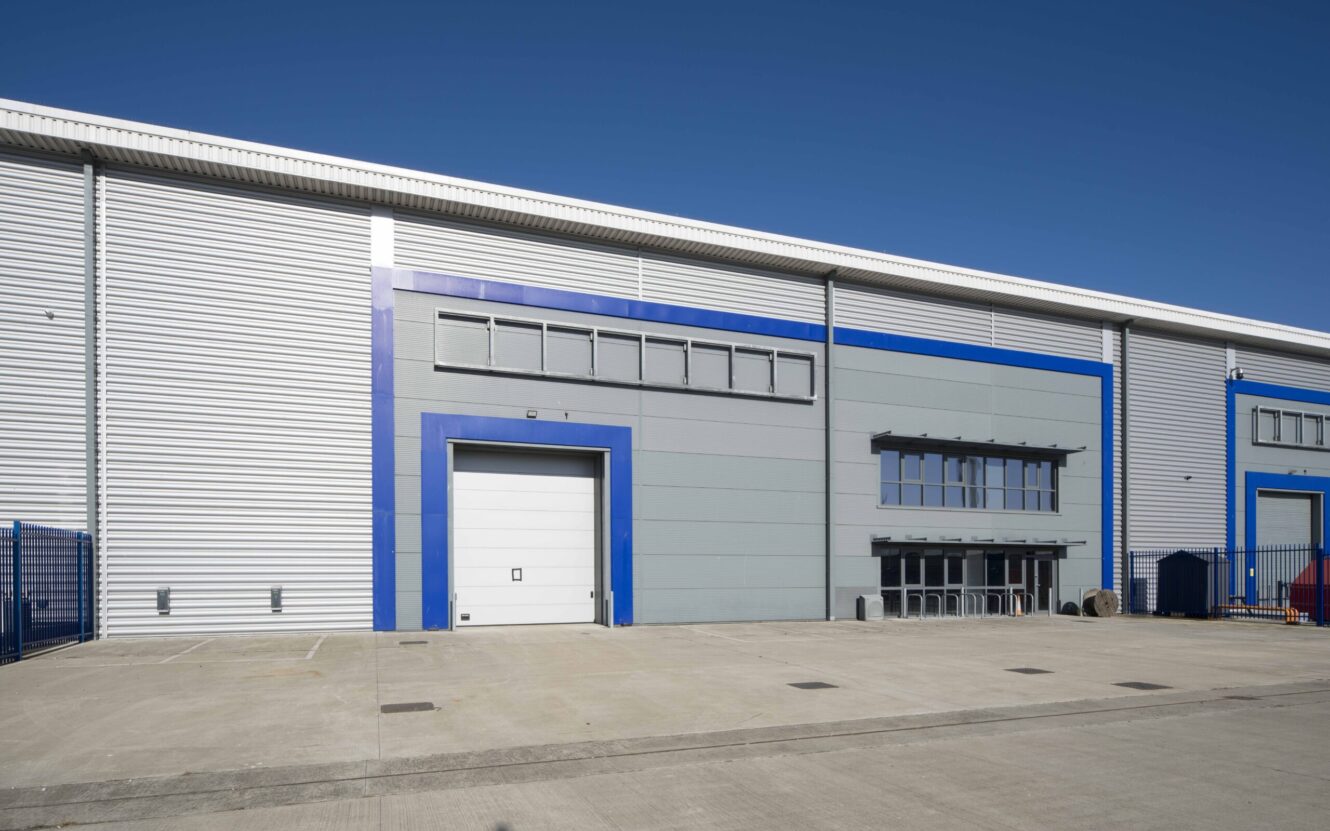 A modern industrial warehouse with a large white loading bay door, blue trim, metal cladding, and several windows on a clear day.