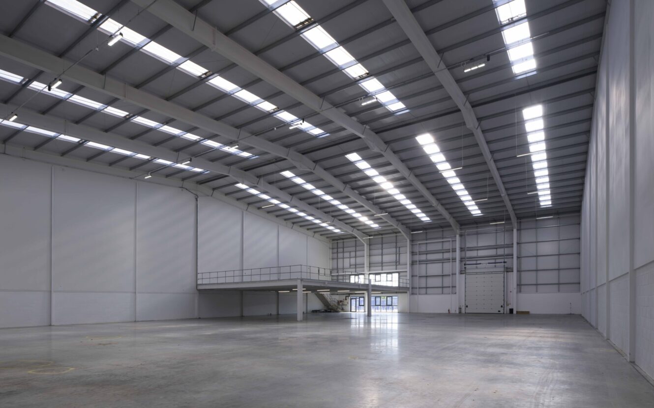 Large, empty industrial warehouse with high ceilings, skylights, white walls, and a mezzanine platform. The floor is concrete and there is a large loading door at the back.